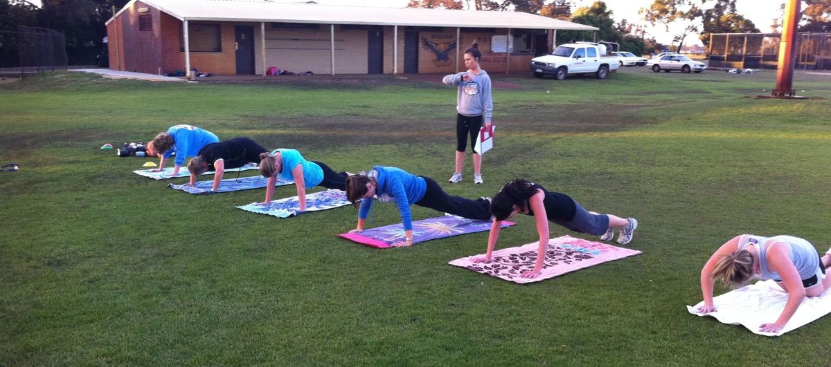 A WA Bootcamps for Women session on a sports oval in Perth: a coach with a clipboard times a line of members holding push-up positions on their towels